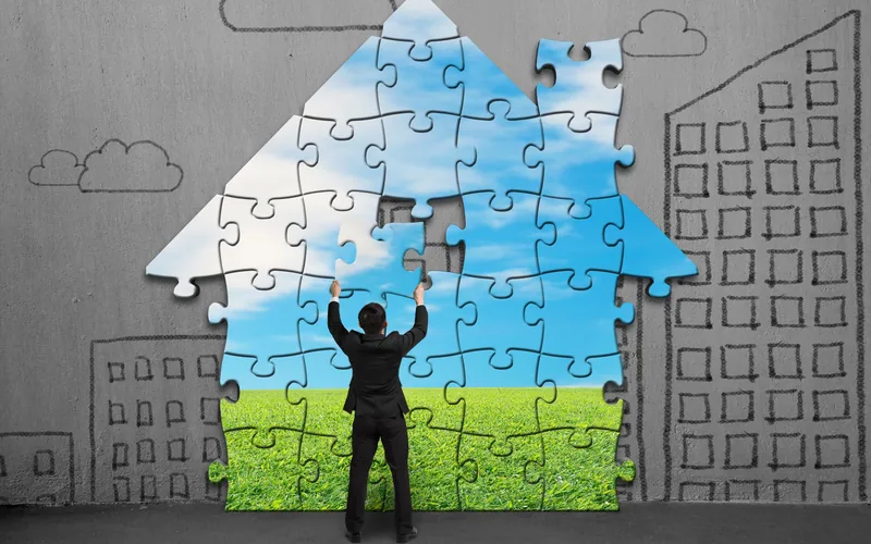 Person setzt ein Haus aus grossen Puzzleteilen zusammen, dahinter an die Wand gezeichnete Häuser.