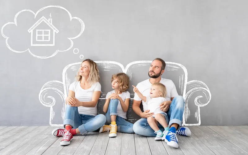 Familie mit zwei Kindern sitzt vor einem an die Wand gezeichneten Sofa, darüber ein Haus in einer Gedankenblase.